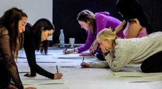 A photograph of a group of people kneeling on the floor drawing on sheets of paper.