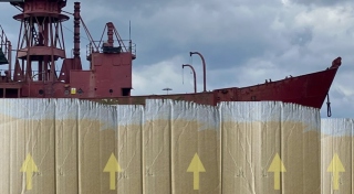 A vertical wall of card board with directional yellow arrows pointing upward sits in the foreground. In the background and behind the cardboard wall, there is a large red ship.