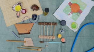 A variety of paint brushes and paints are laid out on a table beside a painting on handmade paper, various seashells and a bamboo mat