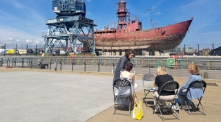 A photograph of seated people drawing in front of a red large ship.