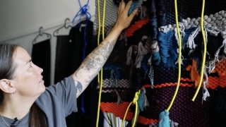 Emily stands profile to camera and is reaching up to touch a grey, blue, red, black and purple woven tapestry with lime green cords.