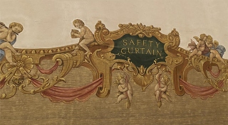 A phone photograph of the painted safety curtain in the Gaiety Theatre, Dublin, which is gold, cream, green and a faded red. Painted chubby cherubs side either side of a baroque frame which reads ‘SAFETY CURTAIN’.
