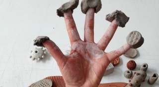 An outstretched hand has chunks of clay playfully pressed into each finder tip. In the background there are slabs of clay placed on a table.
