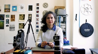 Atoosa sits at a table in her studio facing forward and looks directly at the camera. She has long curly dark hair that is tied in a low ponytail and sits just past her shoulders. She wears a white knit sweatshirt and a blue floral shirt. On the table in front of her are an array of analogue camera parts.