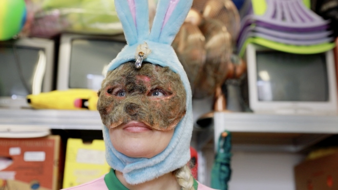Ella Bertilsson wears a blue soft fluffy bunny-rabbit head piece and a brown furry eye-mass. She faces the camera and looks up and to the left left into the distance.