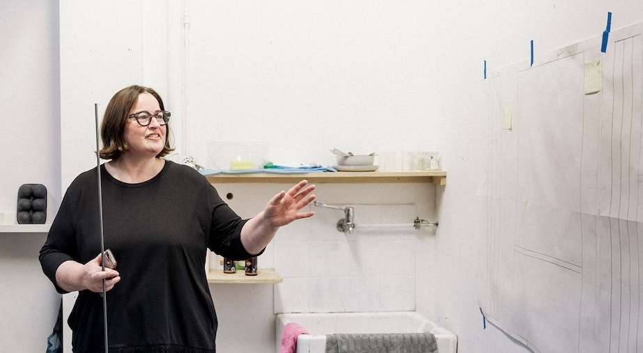 DABF26 Tour: Open Studio with Artist Joanne Reid

Joanne stands in a white studio space, holding a steel rod like a staff, and looks and points at large drawings on white paper stuck to the wall with blue painter's tape.