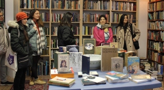 A group of six people looking in various directions stand in front of a wall of bookshelves filled with brightly coloured books. A display of books of varying size and topics is on a small table to the right in front of the group.
