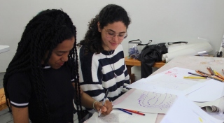 Two people sit at a table at a drawing workshop. The person on the left has a marker in their hand and is dawring on a white piece of paper. The person on the right is watching.