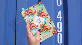 A photo of a hand holding up a book with a colourful cover. In the middle of the cover are the words 'Philip K. Dick, Ubik'. The book is held up in front of the corrugated metal exterior of a shipping container, which is painted blue.