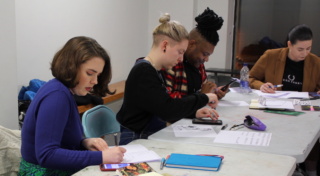 a group of young people are seated at a table in an art gallery. Each person has a piece of paper or notebook and an assortment of pens in front of them and each is referencing imagery on their phones.