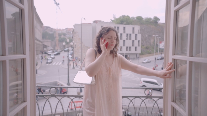 A woman stands facing the camera on a balcony, the street is behind her. She holds a red phone to her ear and her eyes are closed as she leans against the balcony door with her left hand.