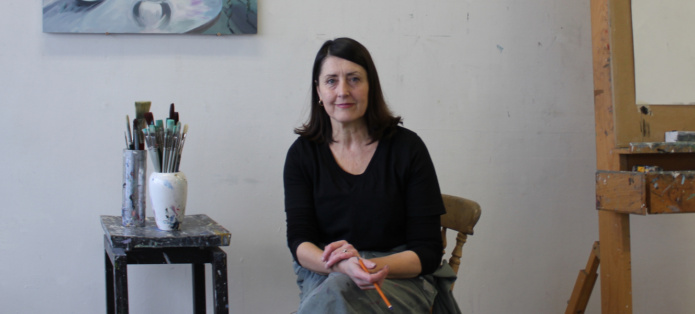 Mairead O'hEocha sits on a chair in her studio. To her right are two jars containing paintbrushes resting on a stool and to her left is an easel, which is cropped out of the frame of the image.