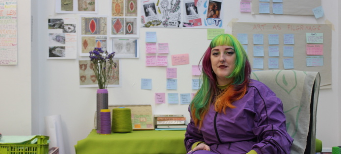 Tara Carrol sits in their studio. They have green pink and orange hair and wear a purple jumpsuit. Behind them an array of image and coloured post its are stuck to the wall.