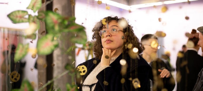 A visitor stands in a gallery space looking up at an artwork made from hanging wires and wristwatch pieces.